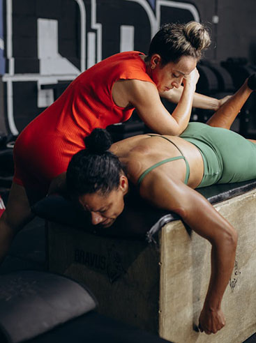 A fitness trainer assists a woman during a massage or recovery session on a gym bench, promoting physical therapy in a workout setting.