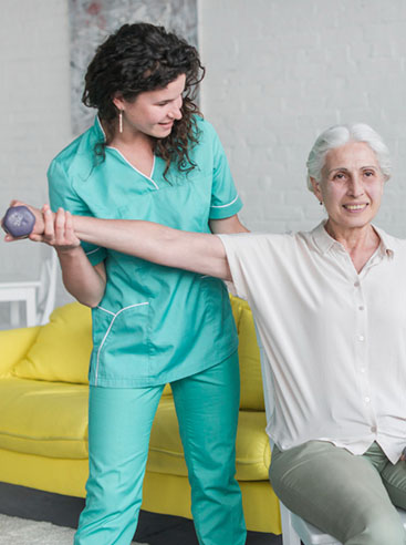 A caregiver assists an elderly woman in exercising with a light dumbbell in a bright, modern room with yellow furniture.